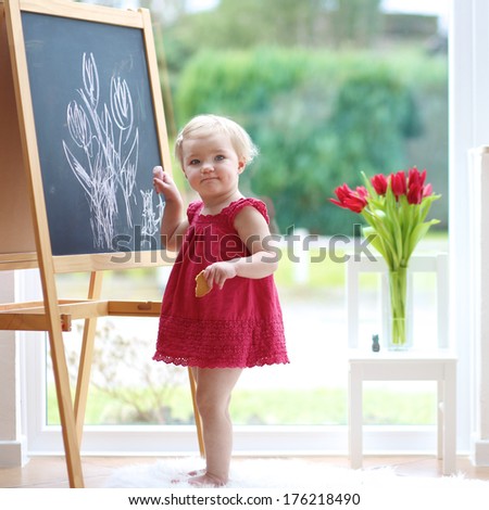 Funny blonde toddler girl in beautiful red dress drawing tulips with chalk on black board standing next to a big window