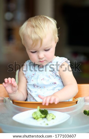 Sweet baby or toddler girl sitting in a high feeding chair biting on delicious freshly cooked broccoli
