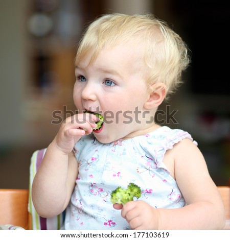 Cute baby or toddler girl sitting in a high feeding chair biting on delicious freshly cooked broccoli
