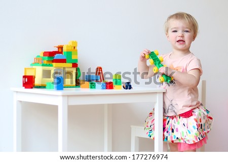 Cheerful toddler girl building house from plastic blocks sitting at small white desk indoors at home or kindergarten