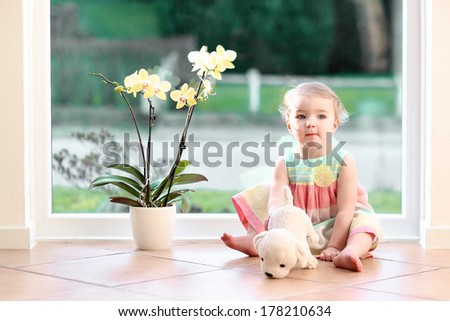 Cute Toddler Girl With Blond Curly Hair Playing Indoors With Puppy Toy Sitting On Tiles Floor In White Sunny Room Next To Big Street View Window With Beautiful Orchid In The Pot