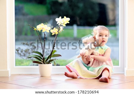 Adorable Toddler Girl With Blond Curly Hair Playing Indoors With Puppy Toy Sitting On Tiles Floor In White Sunny Room Next To Big Street View Window With Beautiful Orchid In The Pot
