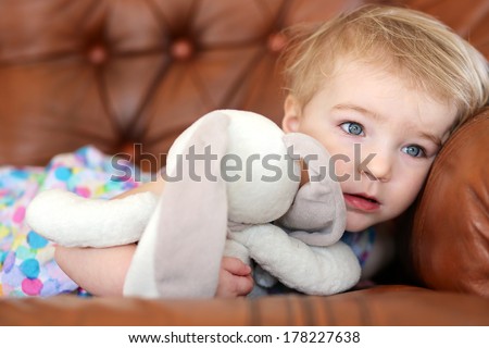 Portrait of cute blonde toddler girl lying on leather sofa holding rabbit toy in her arms