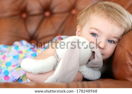 Portrait of adorable blonde toddler girl lying on leather sofa holding rabbit toy in her arms