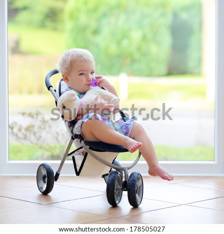 Adorable toddler girl with blond curly hair playing indoors with puppy toy sitting in the doll stroller in white sunny room next to big garden view window