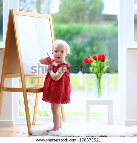 Funny blonde toddler girl in beautiful red dress drawing tulips with crayons on white board standing next to a big street view window