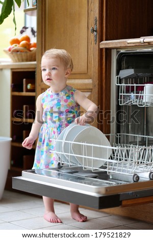 Cute blonde toddler girl helping in the kitchen taking plates out of dish washing machine