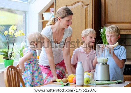 Young mother with three kids, teenager twin sons and little toddler daughter, standing together on sunny kitchen preparing healthy drink with milk and fruits. Boy is placing banana in the mixer.
