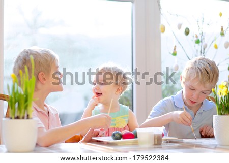 Group of laughing kids from one family, two twin brothers and their little toddler sister, decorating and painting Easter eggs sitting together in the kitchen on a sunny day.