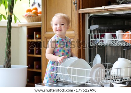 Smiling blonde toddler girl helping in the kitchen taking plates out of dish washing machine