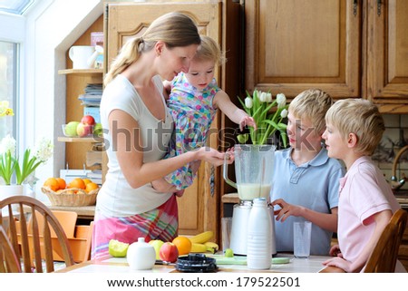 Young mother with three kids, teenager twin sons and little toddler daughter, standing together on sunny kitchen preparing healthy drink with milk and fruits. Girl is placing kiwi in the mixer