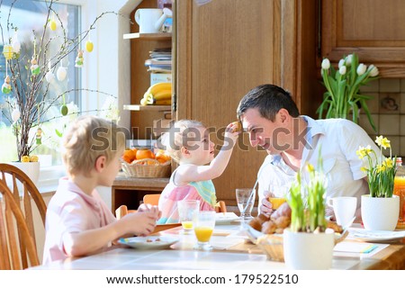 Father with teenager son and toddler daughter eating eggs during family breakfast on Easter day sitting together in sunny kitchen. Selective focus on girl and man.