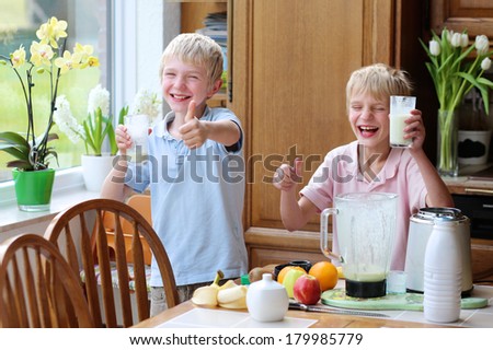Two happy teenager boys, twin brothers, drinking healthy cocktail from milk and fruits standing indoors in sunny kitchen