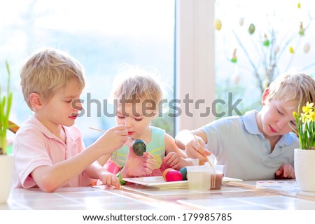 Group of cute children from one family, two twin brothers and their little toddler sister, decorating and painting Easter eggs sitting together in the kitchen on a sunny day. Selective focus on girl.