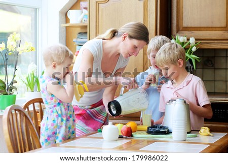 Young mother with three kids, teenager twin sons and little toddler daughter, standing together on sunny kitchen preparing healthy drink with milk and fruits.