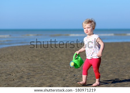 Active blonde little toddler girl in colorful outfit plays with watering can at a shore of the sea on a long calm peaceful beach on a warm sunny summer day