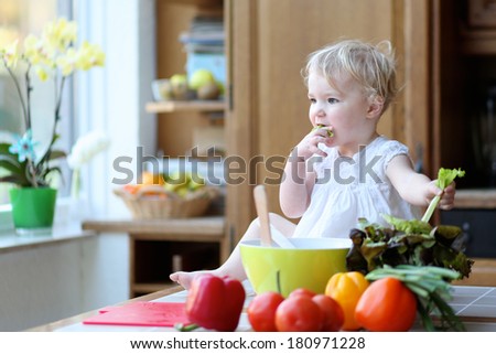 Funny blonde toddler girl in beautiful white dress eating tasty salad leaf preparing healthy vegetables salad sitting on the table in sunny kitchen with big garden view window