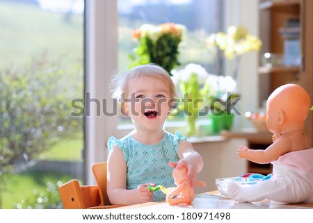 Happy blonde toddler girl feeding with yogurt her dolls sitting on a high chair indoors in sunny kitchen with garden view window