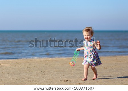 Happy toddler girl in beautiful dress running on the beach on a sunny summer day playing with colorful rainbow spring toy