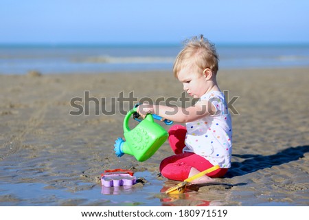 Lovely blonde little toddler girl in colorful outfit plays with watering can at a shore of the sea on a long calm peaceful beach on a warm sunny summer day