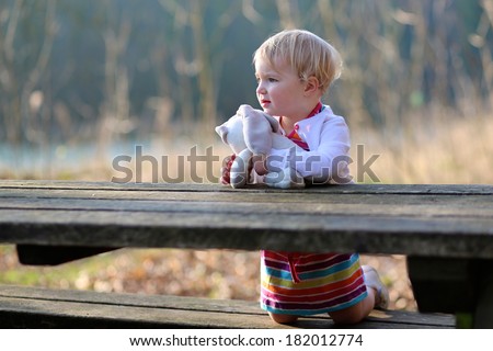 Happy toddler girl with curly blonde hair sitting at picnic table in the forest on a sunny summer day playing with teddy bear toy