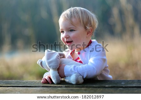 Happy toddler girl with curly blonde hair sitting at picnic table in the forest on a sunny summer day playing with teddy bear toy