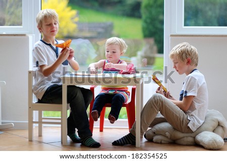 Group of kids, two twin brothers with little toddler sister playing indoors making paper planes sitting nearby big window with street view on a sunny summer day