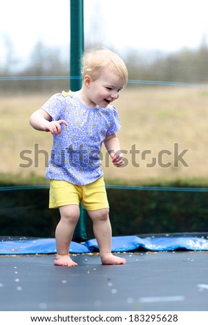Happy blonde toddler girl in beautiful outfit jumping on trampoline in the garden at the backyard of the house on a sunny summer day