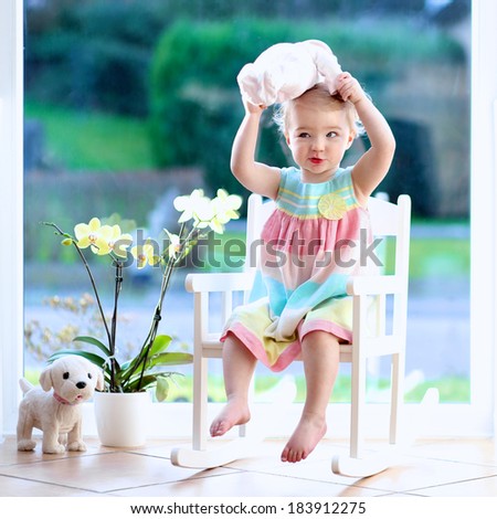 Adorable toddler girl with blond curly hair playing indoors with doll sitting on a rocking chair in white sunny room with big street view window