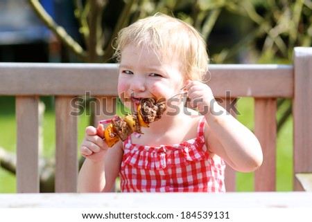 Funny child, adorable blonde toddler girl in red gingnam style dress messy around mouth eating delicious meat made on barbeque sitting outdoors in the garden on a wooden chair on a sunny summer day