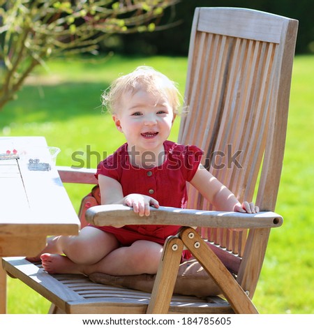 Happy little child, cute blonde toddler girl in beautiful dress snacking outdoors in the garden at the backyard of the house sitting on a wooden teak chair on a sunny summer day eating berries