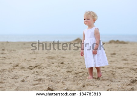 Cute little child, adorable toddler girl in beautiful white dress standing on wet sand on a coastline in the North Sea enjoying vacation on a sunny summer day