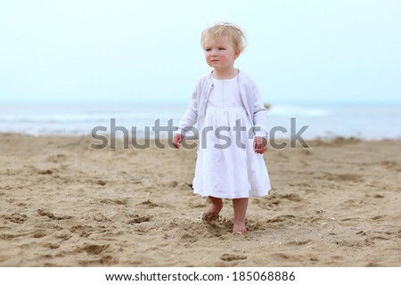 Cute little child, adorable toddler girl in beautiful white dress standing on wet sand on a coastline in the North Sea enjoying vacation on a sunny summer day