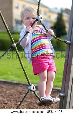 Happy little child, blonde toddler girl in colorful casual suit, having fun on the playground climbing on the net on a sunny summer day