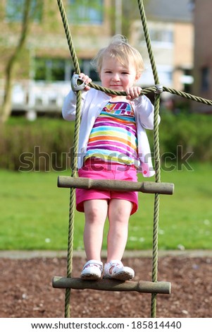 Happy little child, blonde toddler girl in colorful casual suit, having fun on the playground climbing on the rope ladder on a sunny summer day
