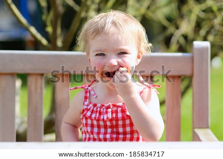 Funny child, adorable blonde toddler girl in red dress messy around mouth eating delicious meat made on bbq sitting outdoors in the garden on a wooden chair on a sunny summer day