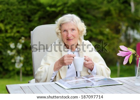 Happy healthy senior woman relaxing in the garden sitting at wooden teak table drinking cup of tea or coffee watching pictures of her baby grandson in family photo album
