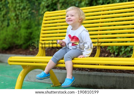Happy little child, adorable toddler girl with blond curly hair in casual outfit enjoying ice cream outdoors sitting on yellow bench in amusement park on a sunny summer day