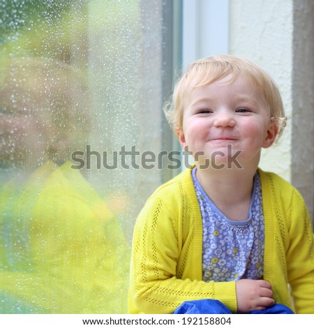 Cute little child, blonde curly toddler girl in colorful casual outfit sitting indoors on a rainy day looking through window with garden view