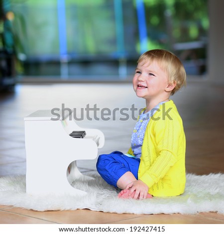 Happy little child, blonde curly toddler girl having fun playing piano toy sitting on the sheepskin lying on the tiles floor in big room with garden view window