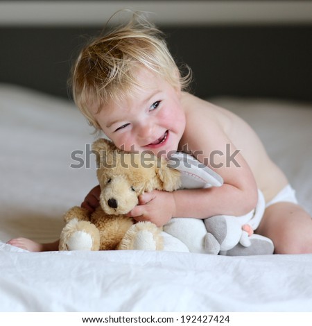 Little sleepy child, just awake blonde curly toddler girl, playing with teddy bear sitting indoors in bedroom on big parents bed on an early sunny morning