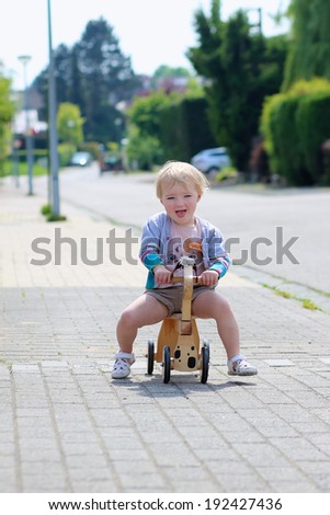 Happy little kid, cute blonde toddler girl playing outdoors on the street riding her push bike, wooden horse with wheels, on a sunny summer day