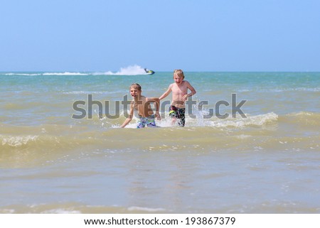 Two happy children, twin brothers, blonde school boys playing on the beach jumping and splashing in the sea on a sunny summer day