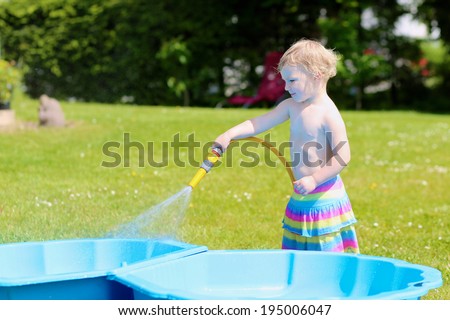 Happy little child, funny blonde toddler girl playing with water hose outdoors in the garden at the backyard of the house on a hot sunny summer day
