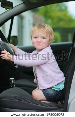 Happy little child, funny blonde toddler girl sitting on her knees inside of the car on driver seat holding steering wheel