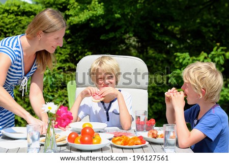 Happy family, mother with kids, two funny schoolboys, laughing and having healthy lunch or dinner outside sitting at picnic table in the garden at the backyard of the house on a sunny summer day