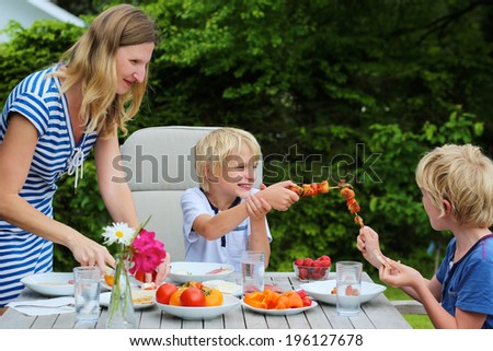 Happy family, mother with kids, two funny schoolboys, laughing and having healthy lunch or dinner outside sitting at picnic table in the garden at the backyard of the house on a sunny summer day