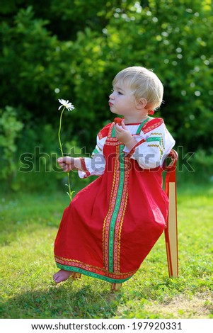 Happy little child, blonde curly toddler girl in traditional russian dress playing outdoors in the garden eating fresh strawberry and smelling daisy flower sitting on small wooden chair