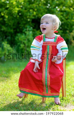 Happy little child, blonde curly toddler girl in traditional russian dress playing outdoors in the garden sitting on small wooden chair