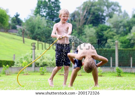 Happy laughing children, two young school boys, enjoying hot sunny summer vacation day playing outdoors in garden at the backyard of the house running on green lawn and spraying water from the hose